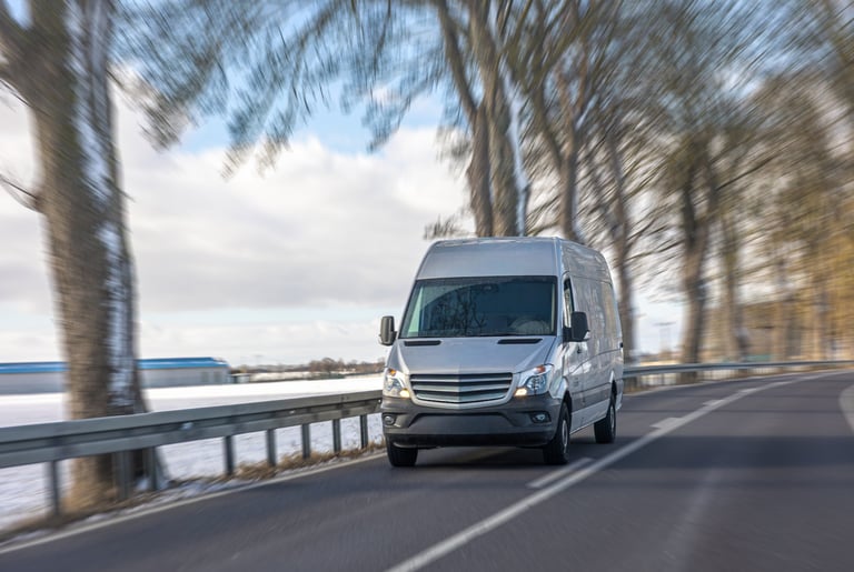 No people cargo van driving on asphalt road along snowy field and trees in daylight. Spacious composition suitable for business, delivery and transportation service concepts